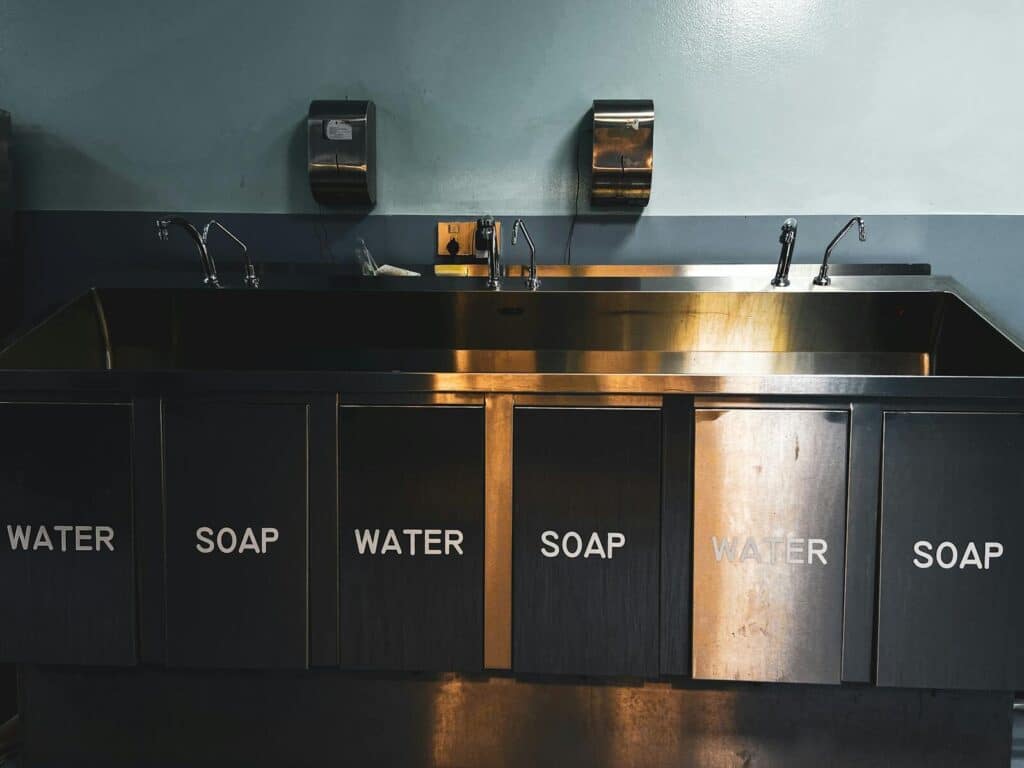 A stainless steel industrial kitchen sink with labeled compartments for water and soap.